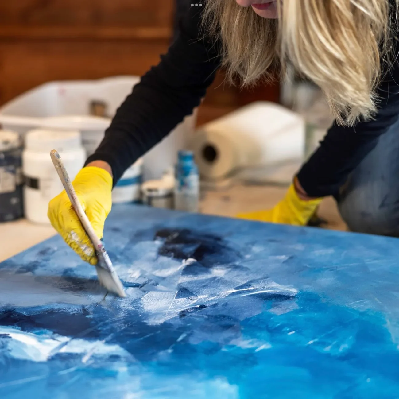 Close-up of artist Rita Thorp’s hand in a yellow glove using a paintbrush to apply thick blue and white paint strokes to a textured abstract painting