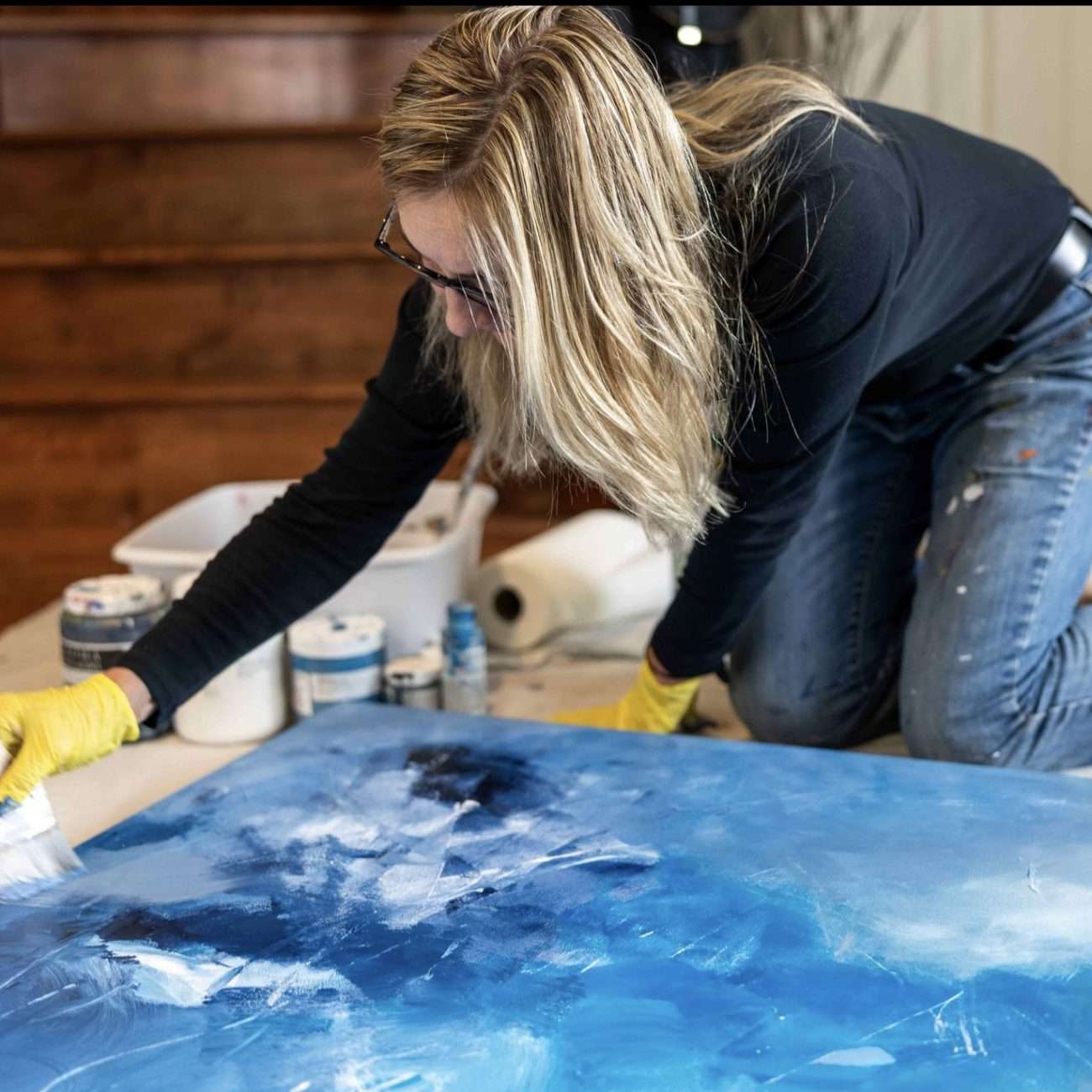 Rita Thorp Canadian Contemporary Artist painting a large abstract blue and white canvas, leaning over while wearing yellow gloves, with various paint supplies in the background.