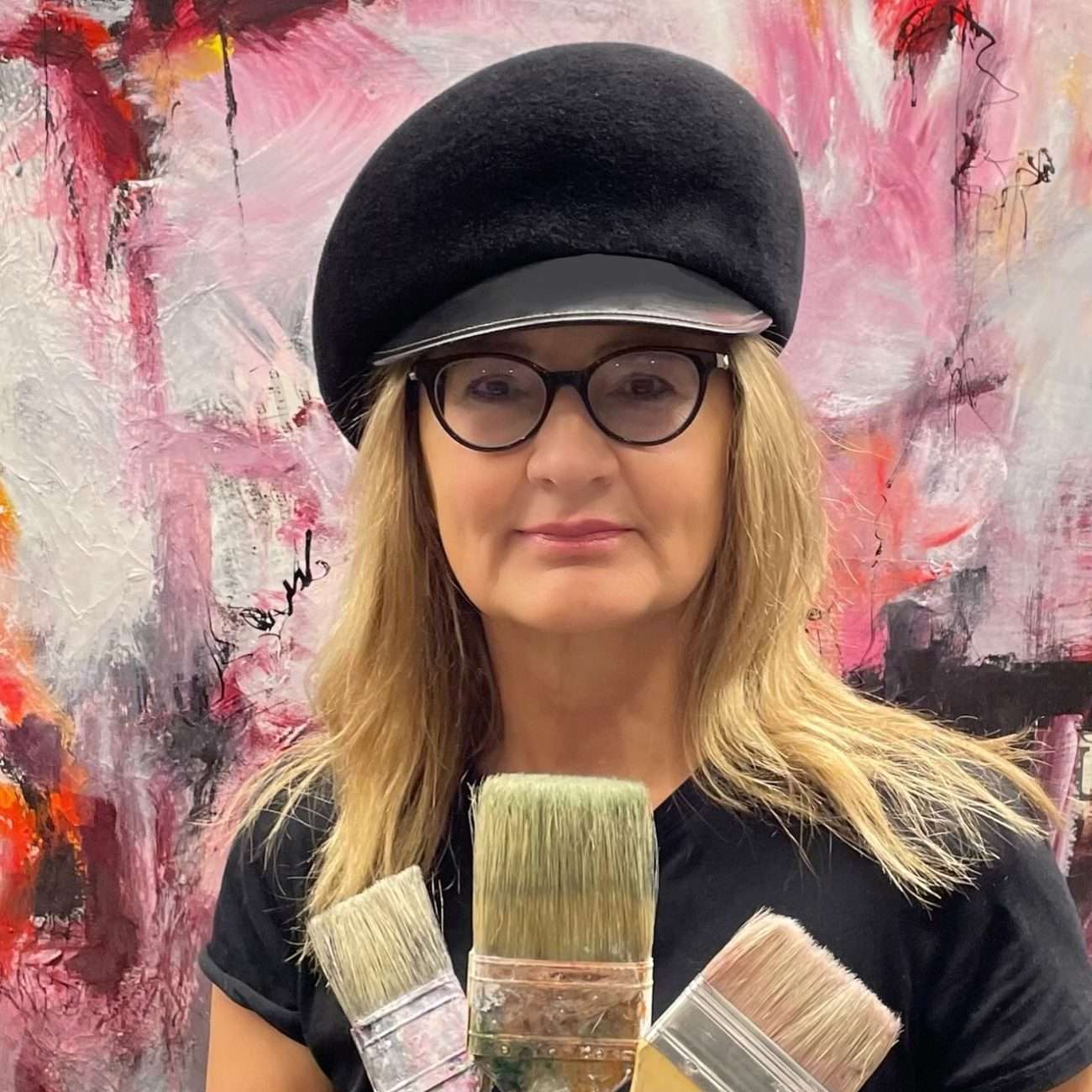 Portrait of contemporary artist Rita Thorp wearing a black beret and glasses, holding multiple paintbrushes in front of a vibrant abstract painting with pink, red, and black tones.